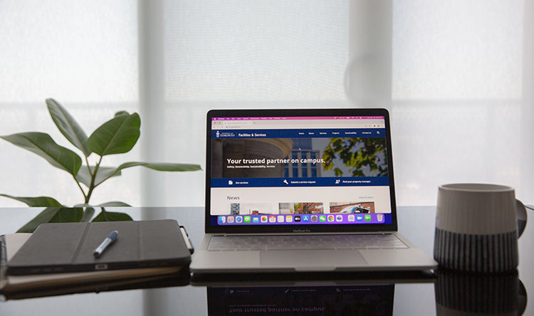 A desk with a coffee mug, plant, notebook, pen, and laptop open to the F&S website.