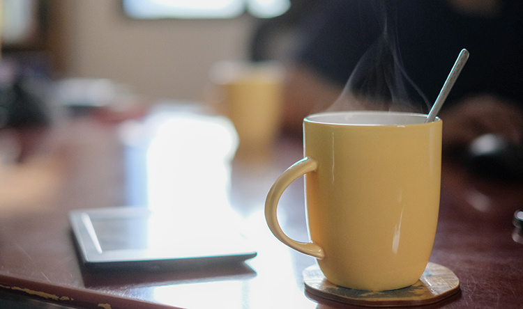 A mug of hot coffee sits on a coaster on a table with a spoon inside.