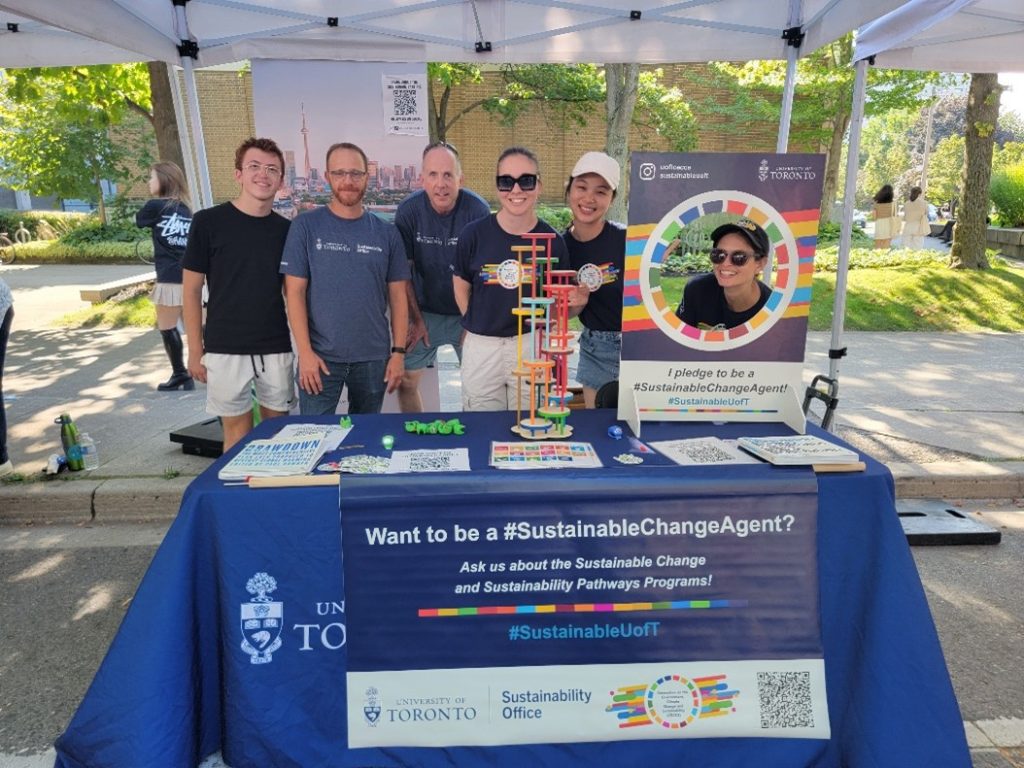 A group of student sustainable ambassadors stand behind a branded table with promotional materials, outdoors on a sunny day.