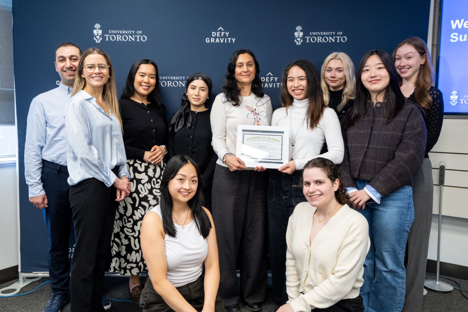 A group photo containing 11 persons. Two of them are jointly holding up a Sustainable Action Award framed certificate. They are standing in front of a U of T blue backdrop that has the crest and "Defy Gravity" repeated as a pattern on it.