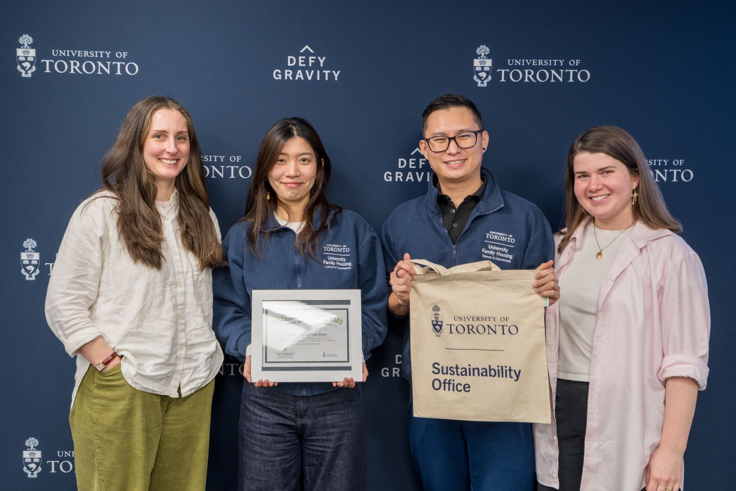 Four people lined up in a U of T blue backdrop that has the crest and "Defy Gravity" repeated as a pattern on it. One person holds up a framed certificate that says "Sustainable Action Award" on it, and another holds up a tote bag that says "University of Toronto Sustainability Office" on it. 