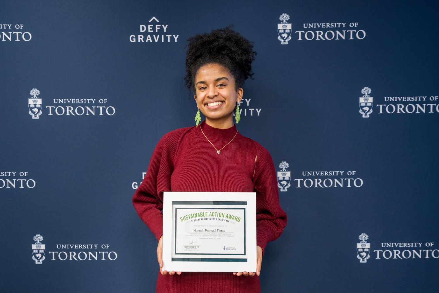 A photo of Hannah holding up her framed Sustainable Action Award certificate. She is smiling and standing in front of a U of T blue backdrop that has the crest and "Defy Gravity" repeated as a pattern on it. 